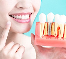 person pointing to their smile and holding a model of a dental implant in the jaw